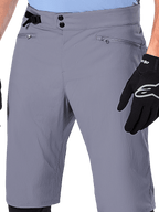 Alpinestars A-Dura Shorts, MTB-shorts, mørkegrå, nærbillede af taljen med dobbelt trykknaplukning, sidejustering af taljen og lynlåslommer, kombineret med sorte cykelhandsker, holdbart mountainbike-tøj