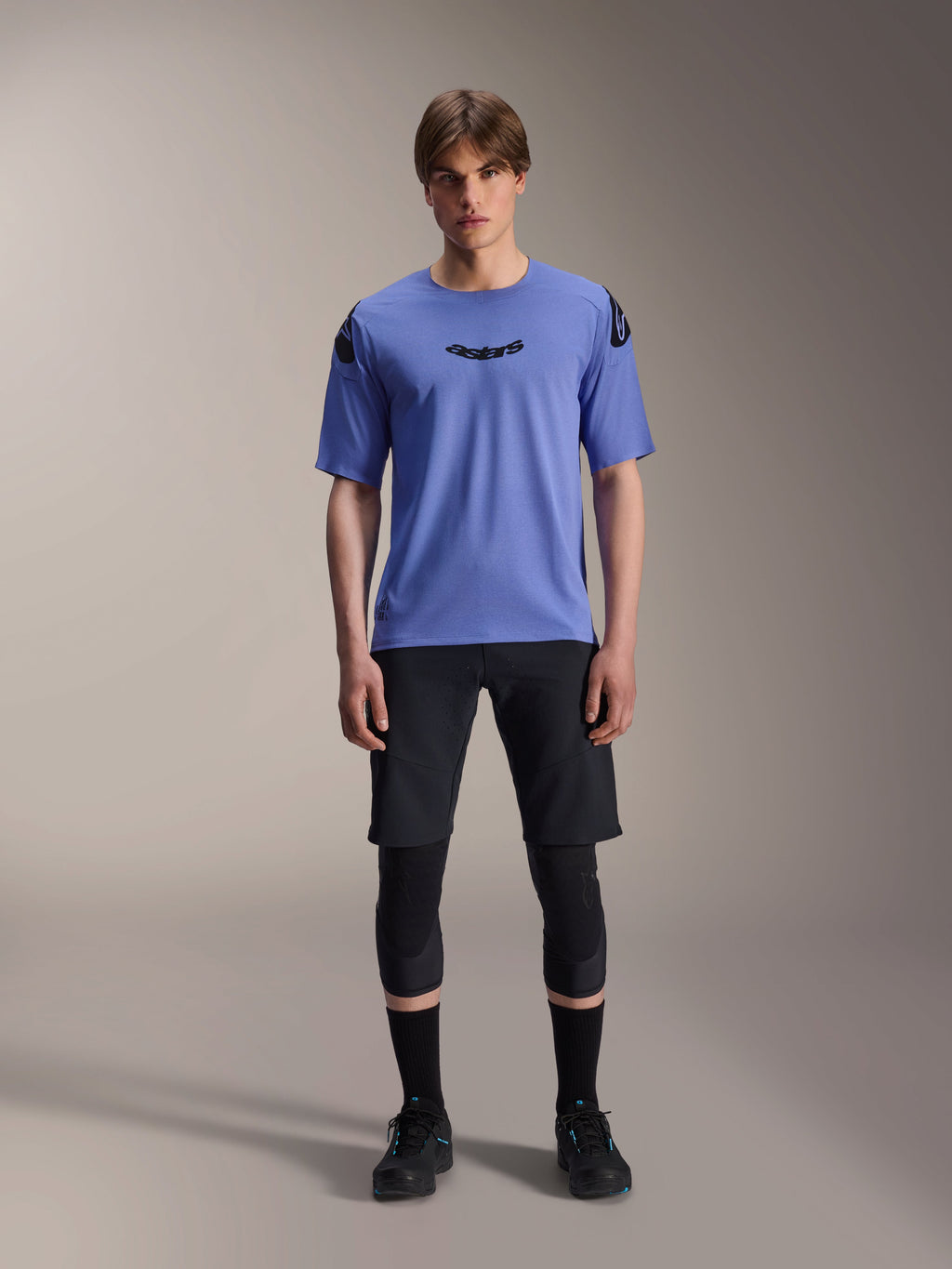 Mandlig model iført Alpinestars A-Aria Polartec Elite kortærmet jersey i Baja Blue, sort astars-logo på brystet, sorte MTB-shorts, knæbeskyttere og sorte cykelsko, studiefoto af mountainbike-udstyr