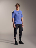 Mandlig model iført Alpinestars A-Aria Lite-shorts i sort, kombineret med en blå A-Aria Polartec Elite kortærmet trøje, sorte knæbeskyttere og sorte cykelsko, performance mountainbike-beklædning til trail-kørsel.