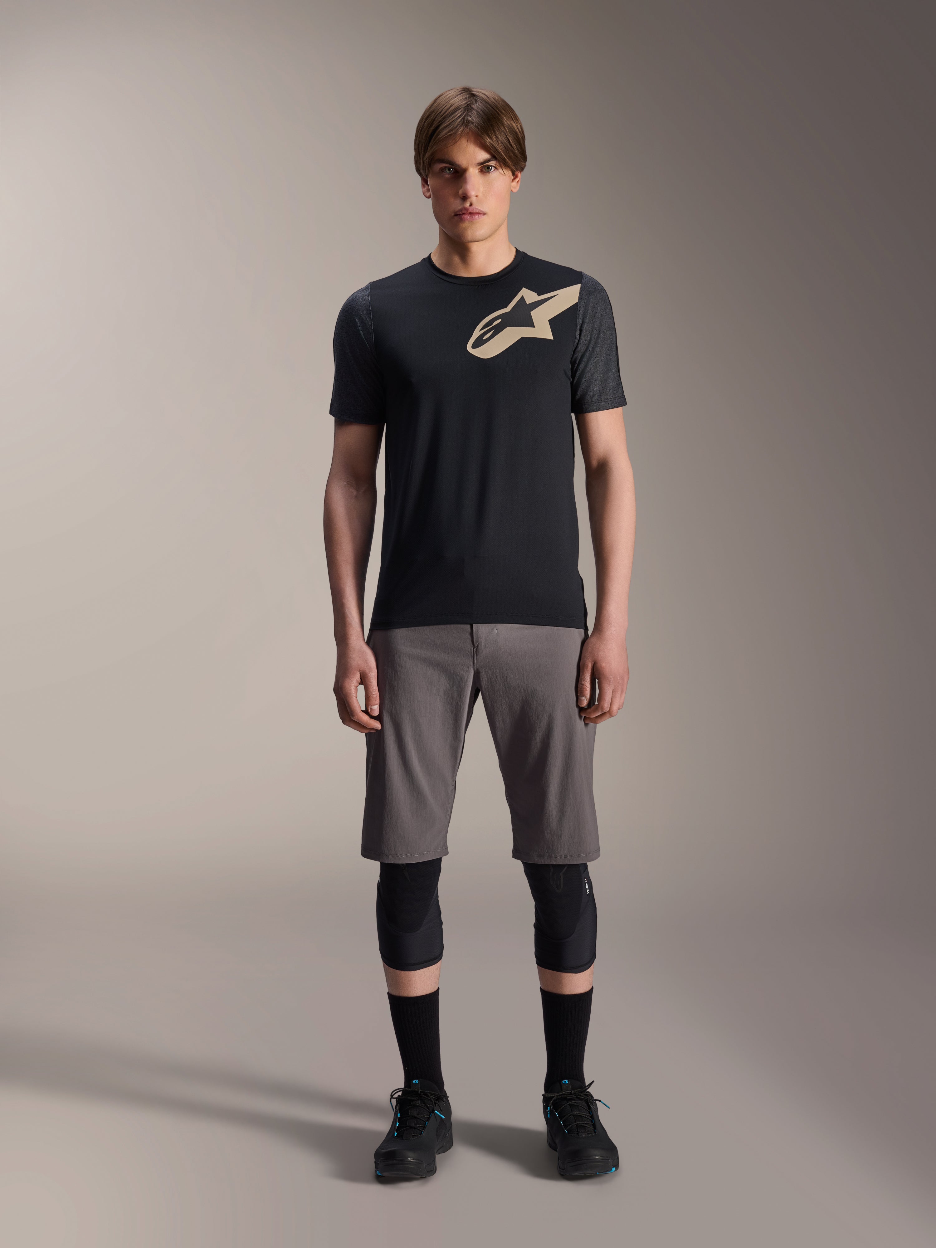 Alpinestars A-Dura Lipan kortærmet jersey, sort med gråmêlerede ærmer og tan-farvet Astars-logo på brystet, båret af en mandlig model med grå mountainbike-shorts og sorte knæbeskyttere, performance MTB-køreudstyr