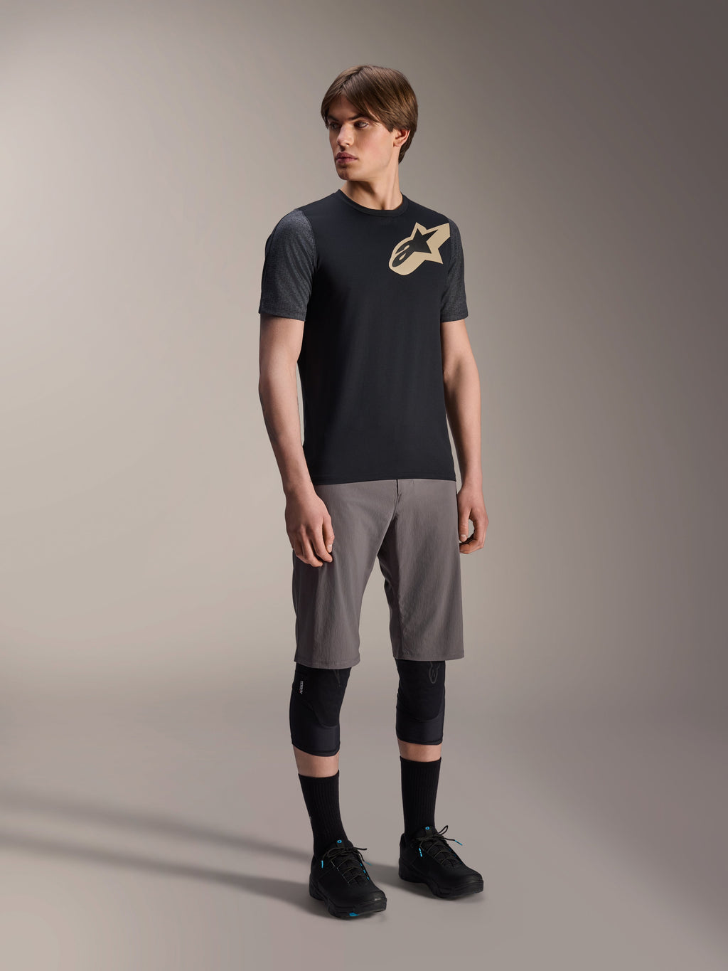 Alpinestars A-Dura Lipan kortærmet jersey i sort, mørkegrå ærmer og et tan Astars-logo på brystet, kombineret med grå mountainbike-shorts, sorte knæbeskyttere og sorte cykelsko, koordineret mountainbike-udstyr