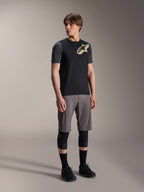 Alpinestars A-Dura Lipan kortærmet jersey i sort, mørkegrå ærmer og et tan Astars-logo på brystet, kombineret med grå mountainbike-shorts, sorte knæbeskyttere og sorte cykelsko, koordineret mountainbike-udstyr