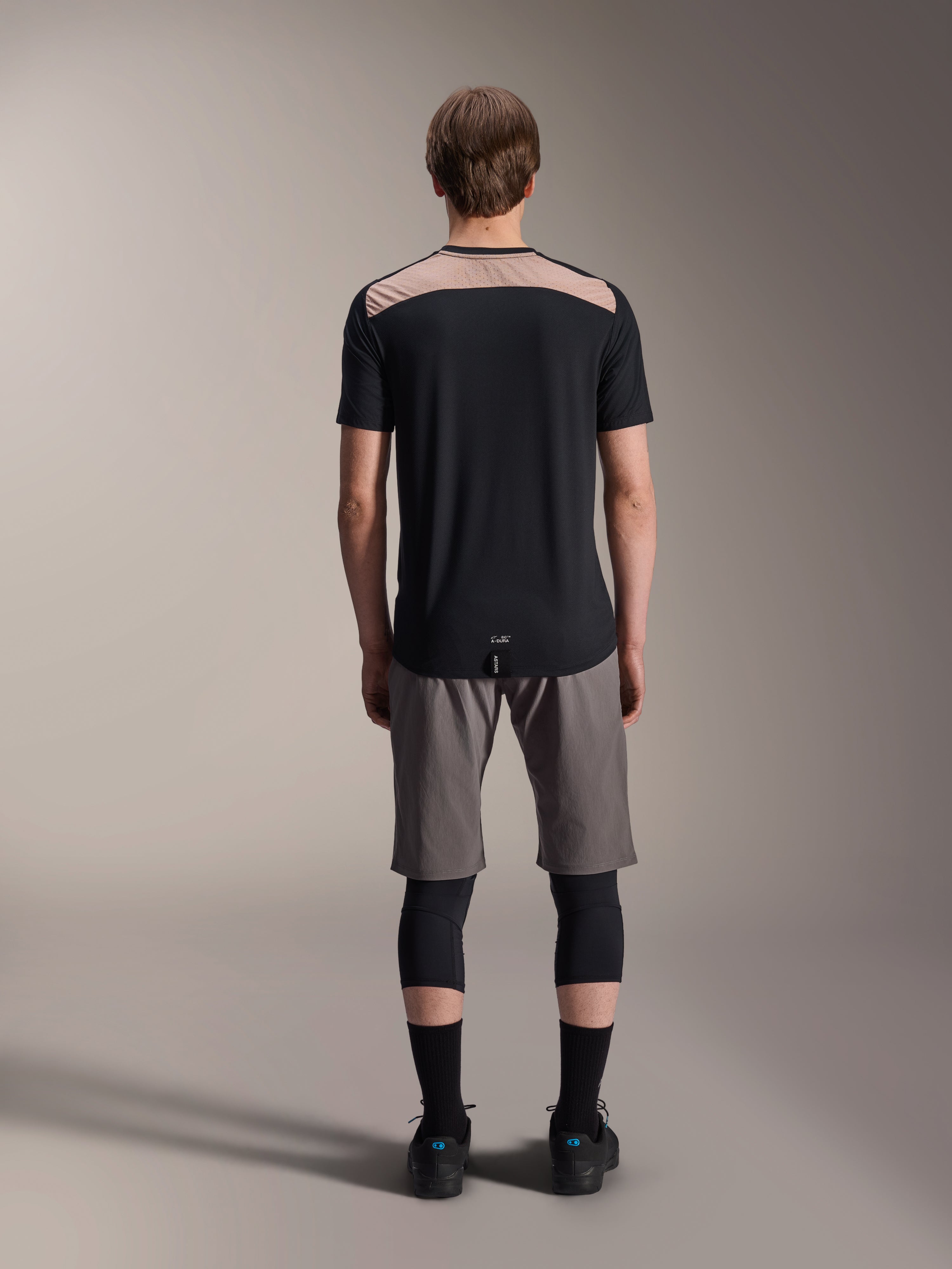 Alpinestars A-Dura Lipan Jersey - Kortærmet, sort med lysebrunt øvre rygpanel, set bagfra med forlænget rygstykke, båret med grå MTB-shorts og sorte knæbeskyttere, performance mountainbike-beklædning