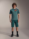 Alpinestars A-Dura Shorts i Dark Pine, mountainbikeshorts til mænd, vist på en model med en matchende blågrøn kortærmet jersey med rødt og sort stjernelogo, sorte sokker og cykelsko, afslappet sportsligt mountainbike-tøj