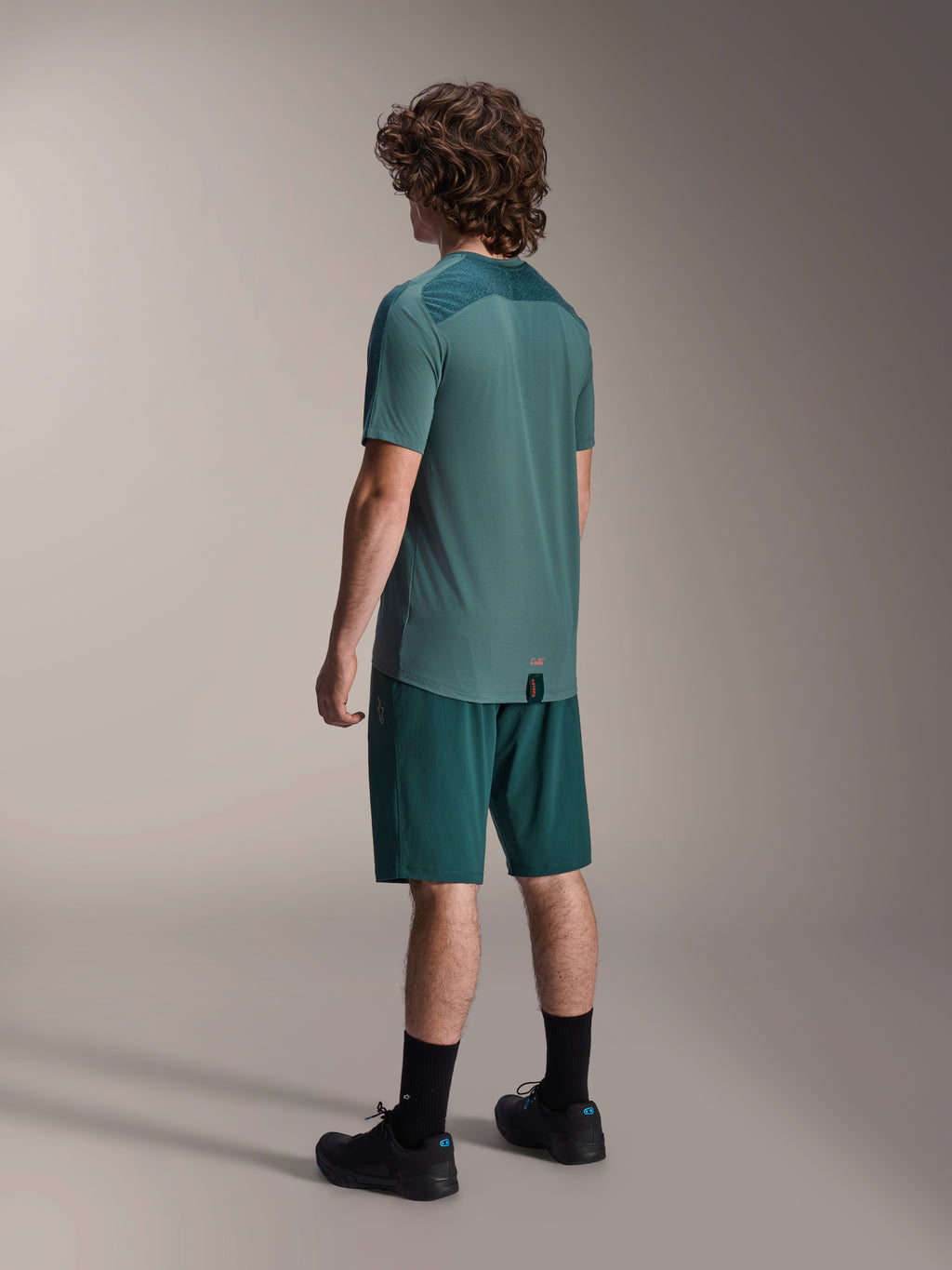Model iført Alpinestars A-Dura shorts i Dark Pine og matchende grøn Lipan kortærmet jersey, set bagfra, kombineret med sorte sokker og sorte mountainbike-sko, professionelt MTB-køreudstyr.