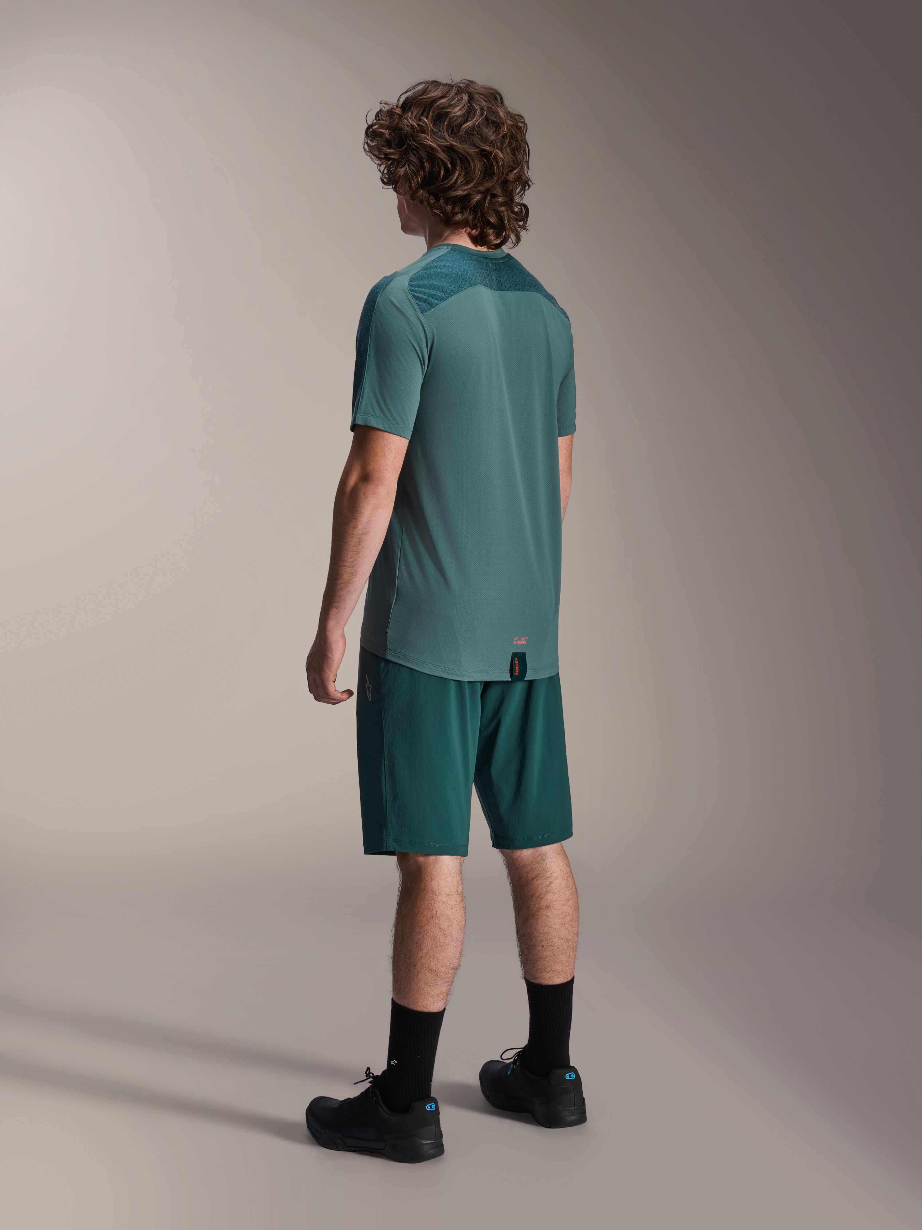 Mandlig model iført Alpinestars A-Dura Lipan kortærmet jersey i Silver Pine, set bagfra, kombineret med matchende mørkegrønne mountainbike-shorts, sorte strømper og sko, med et tekstureret øvre rygpanel og et forlænget bagstykke for ekstra dækning.