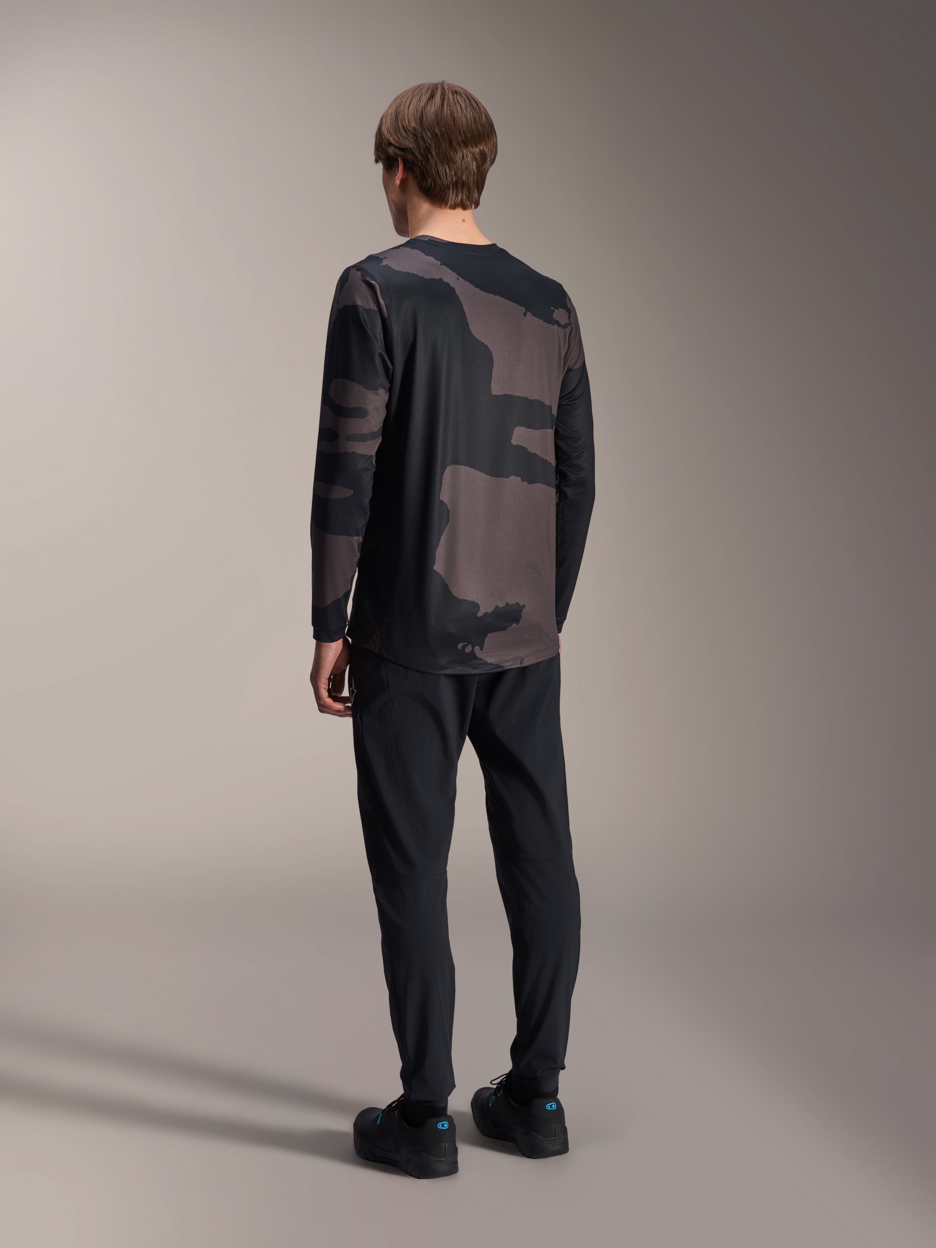Alpinestars A-Dura Hollow Camo langærmet jersey, Black Camo, model set bagfra, teknisk mountainbike-jersey med forlænget ryg, kombineret med sorte bukser og cykelsko, afslappet off-road performance-udstyr