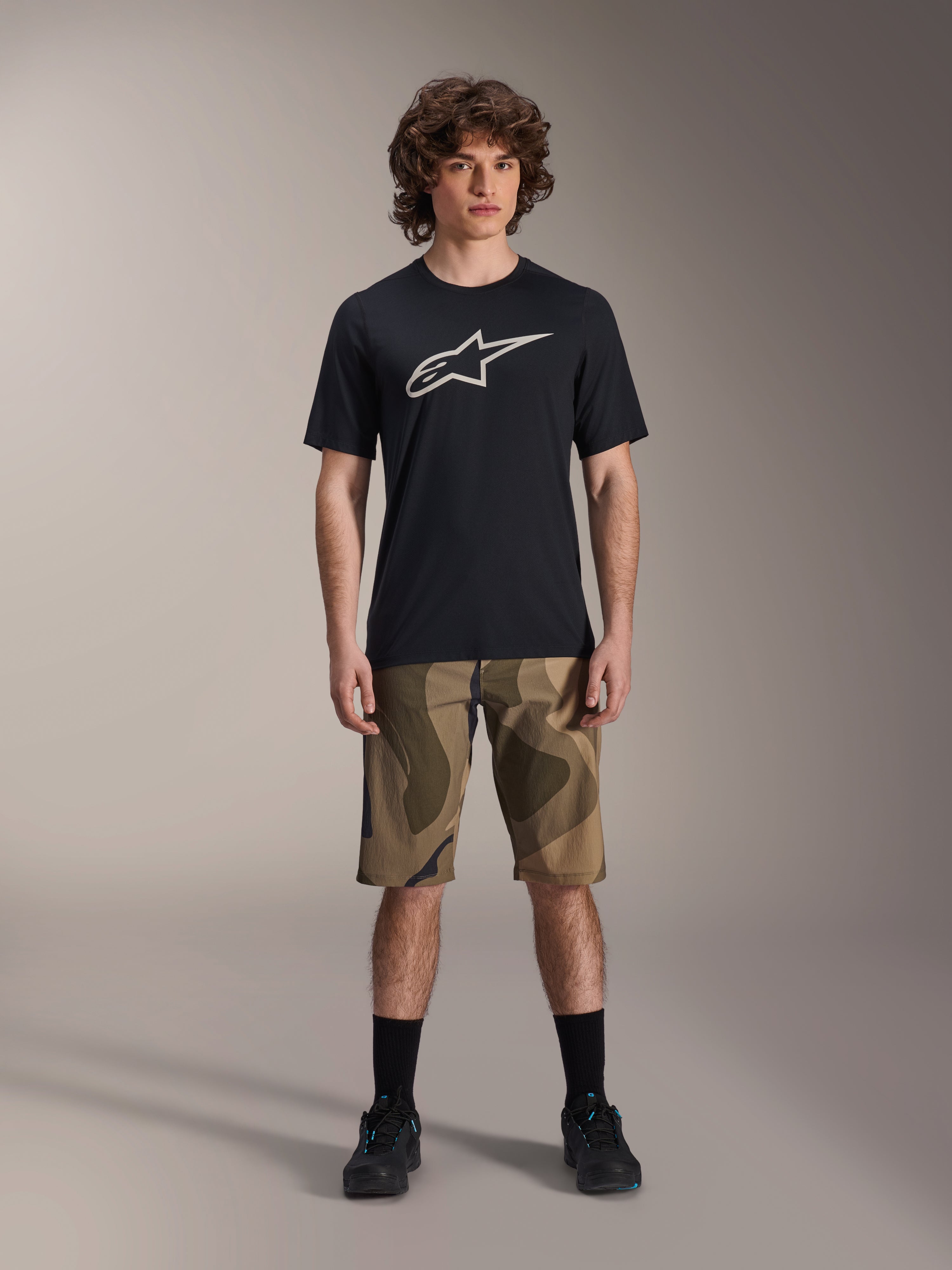Alpinestars A-Dura Astar kortærmet jersey, sort med stort hvidt Astars-logo på brystet, båret af en model med camouflage MTB-shorts og sorte cykelsko, afslappet mountainbike-beklædning.