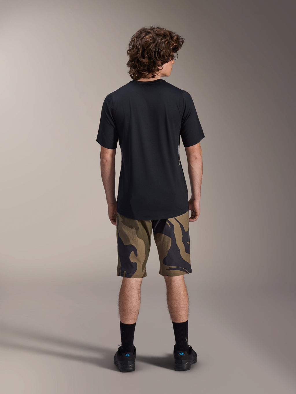 Alpinestars A-Dura Astar kortærmet trøje, sort, set bagfra på en model kombineret med MTB-shorts i camouflagemønster og sort fodtøj, afslappet teknisk mountainbike-beklædning