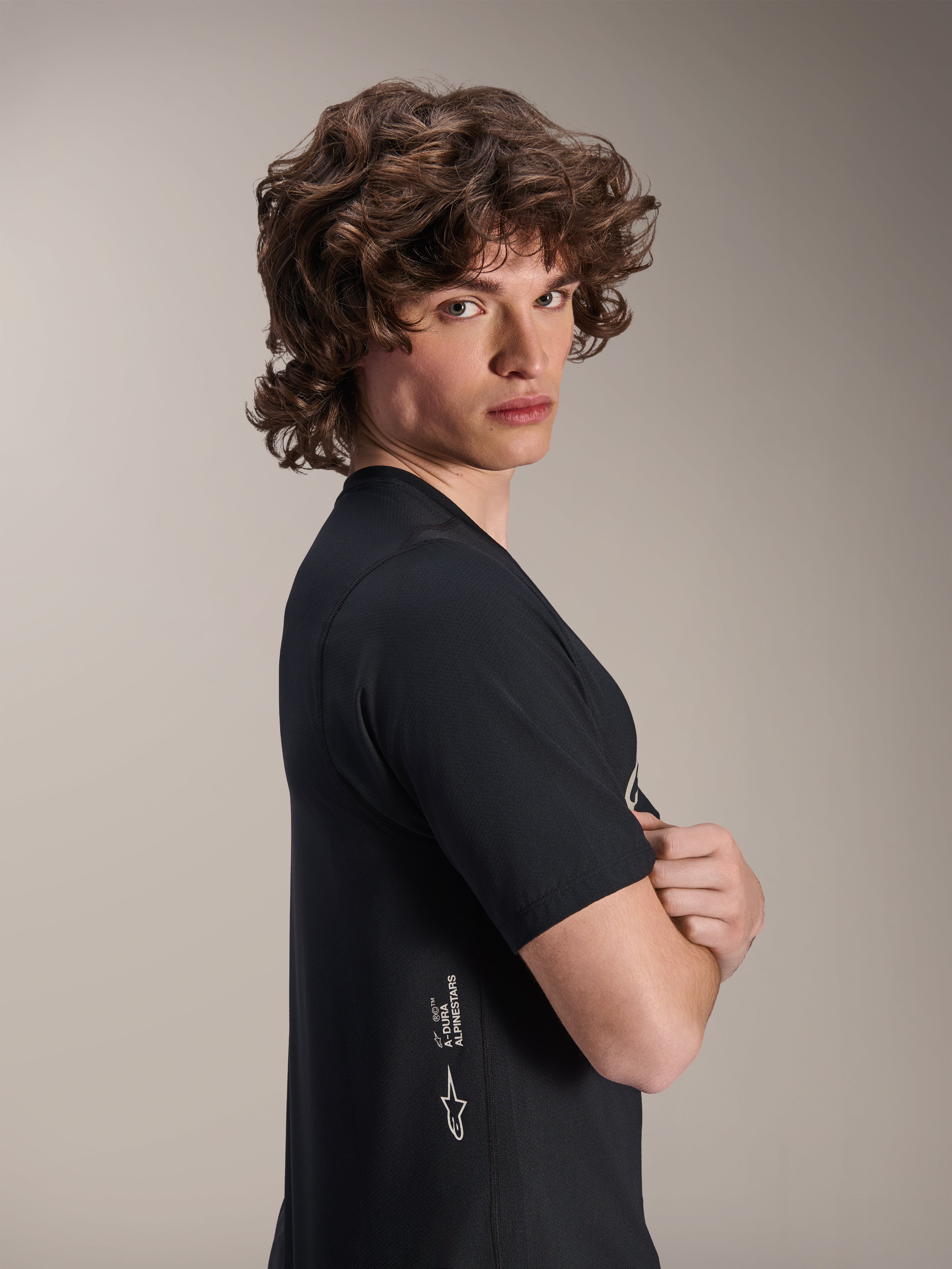 Alpinestars A-Dura Astar Jersey, kortærmet MTB-trøje, sort, mandlig model der poserer i profil med korslagte arme, med hvidt Alpinestars-branding på den nederste side, afslappet atletisk design til mountainbiking.