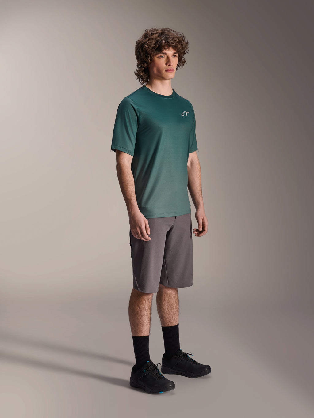 Alpinestars A-Dura Rise Jersey - kortærmet i Silver Pine, grøn mountainbike-jersey båret af en model med grå shorts og sorte sneakers, afslappet MTB-udstyr til trailkørsel, lille hvidt logo på brystet