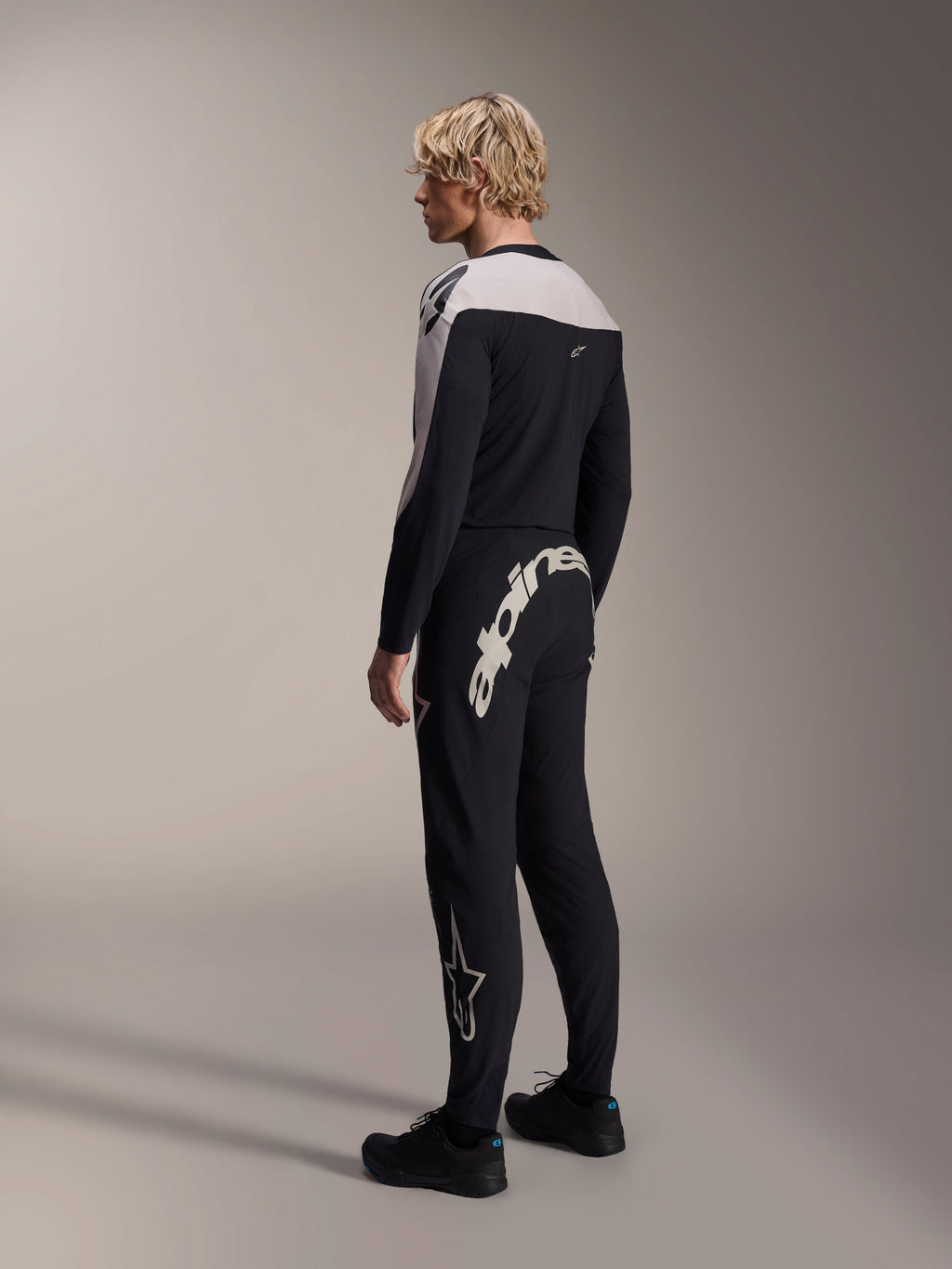 Alpinestars A-Supra Melt Long Sleeve Jersey, Black with white shoulder panels, worn by a model from the back and side, paired with black mountain bike pants featuring a large white wordmark, performance MTB racing kit