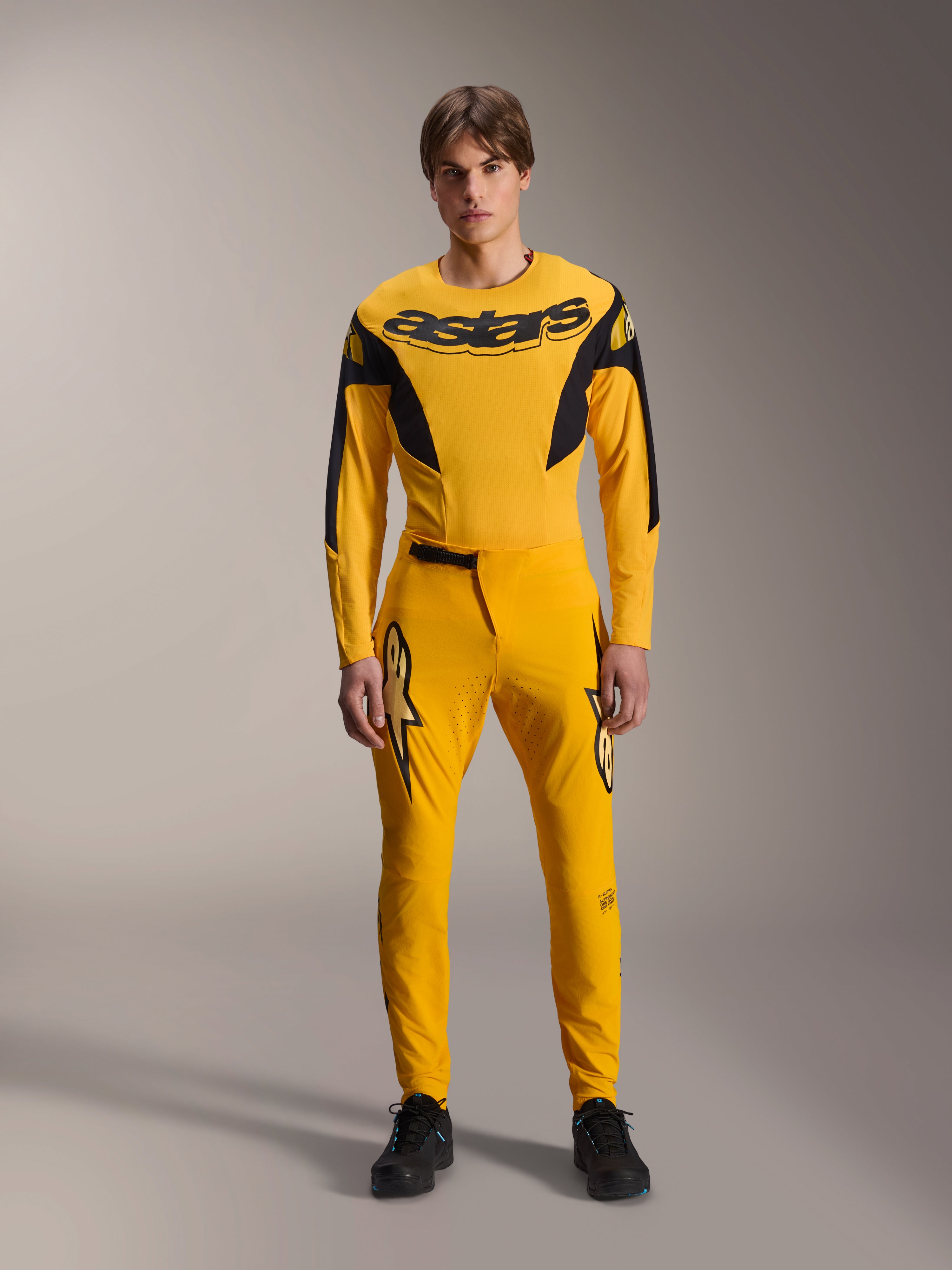Limited Edition A-Supra Melt Gold udstyrssæt, Alpinestars mountainbike-beklædning, mørkegul og sort, model iført langærmet trøje med markant 'astars' brystlogo og matchende performance-bukser, med laserskåret ventilation på inderlårene og en sikker skraldelukning i taljen, professionelt MTB-racersæt