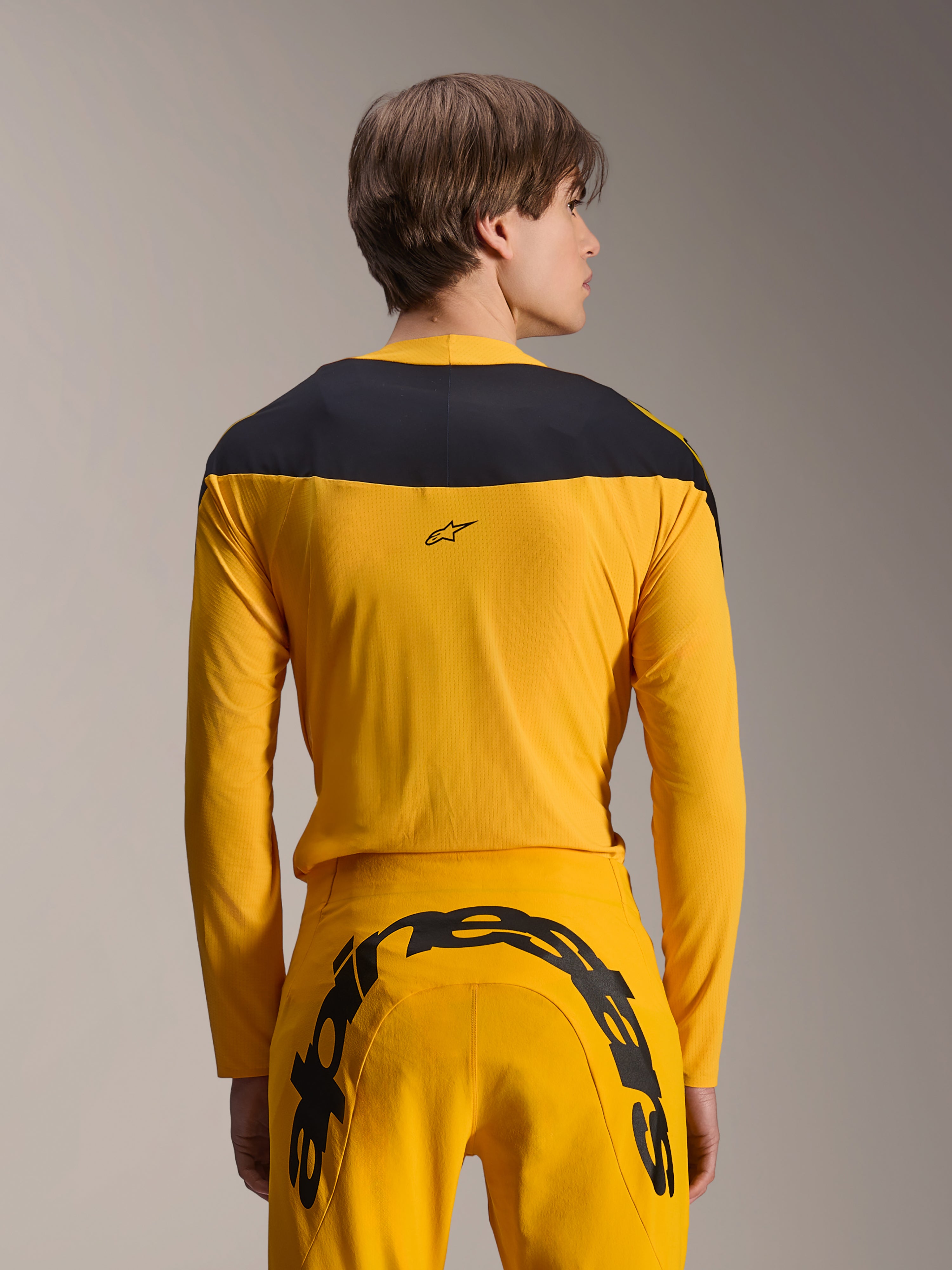 Alpinestars Limited Edition A-Supra Melt Gold Gear-Set, Yellow MTB Gear Set, back view of model wearing long-sleeve jersey with black shoulder panel and matching yellow pants featuring a large black logo on the rear, high-performance cycling apparel.