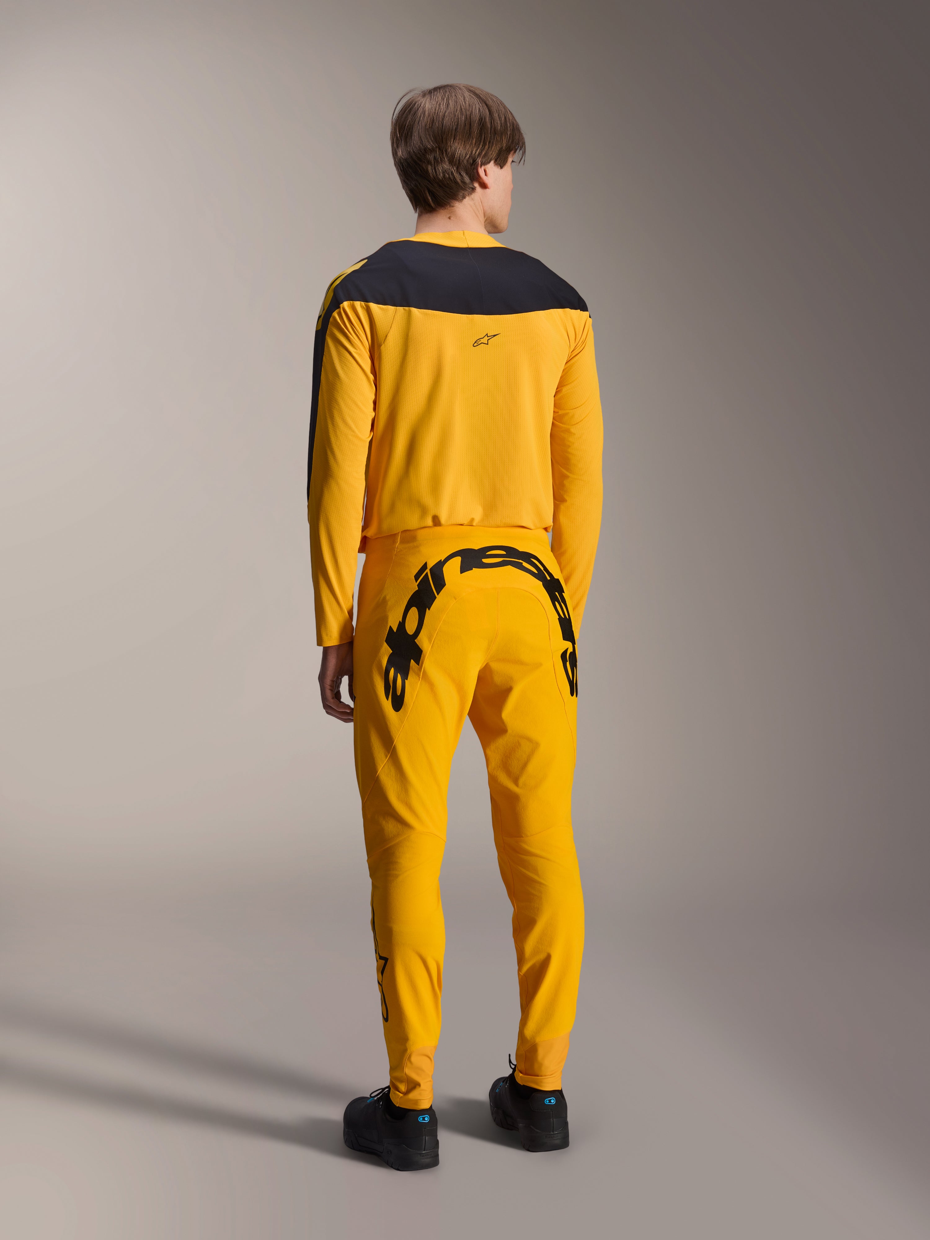 Alpinestars Limited Edition A-Supra Melt Gold-udstyrssæt, MTB-trøje og bukser, mørkegul og sort, set bagfra med stort Alpinestars-navnetræk på bagstykket og sort bærestykke på trøjen, performance mountainbike-tøj