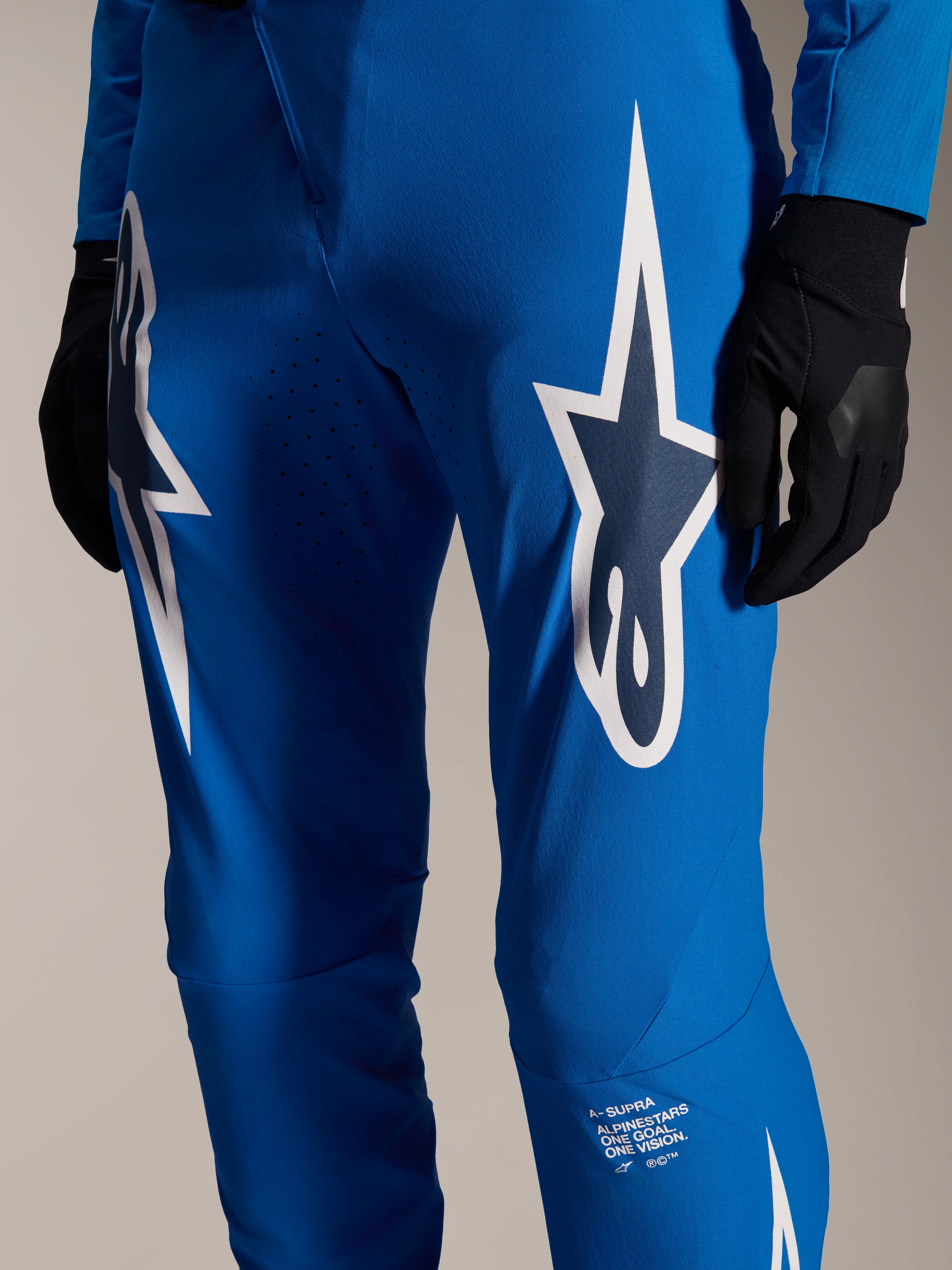 Alpinestars A-Supra Melt bukser, blå cykelbukser, med store hvide Astars-logoer på lårene, laserskårne perforeringer for luftgennemstrømning og signaturbranding på underbenet, bæres med sorte handsker, performance MTB-udstyr