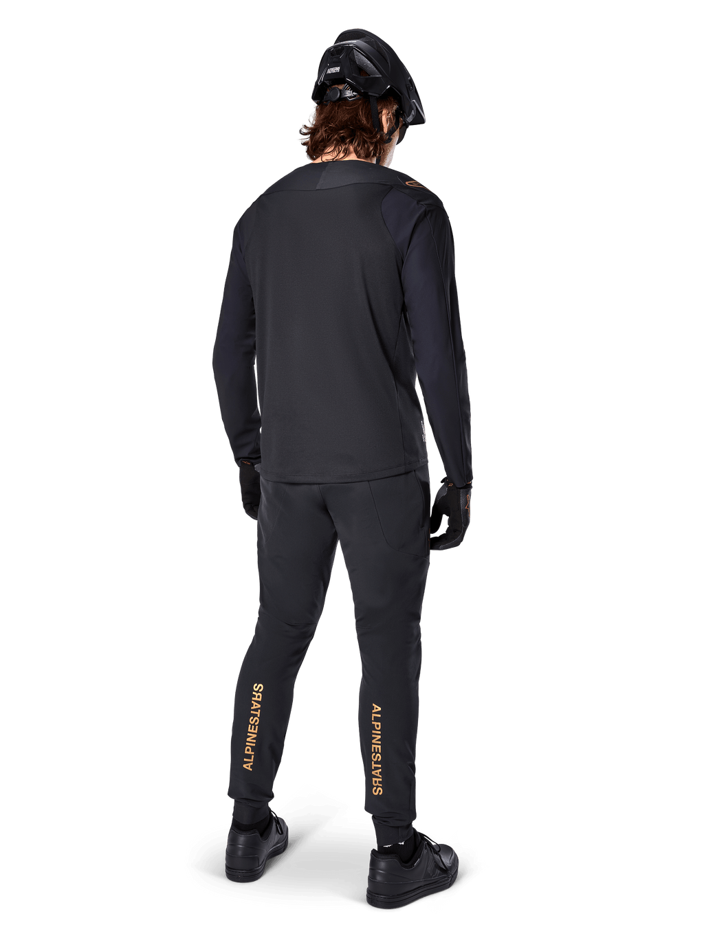Set bagfra af en mountainbike-rytter iført Alpinestars A-Aria Polartec Switch langærmet jersey i sort, kombineret med sorte MTB-bukser med tan-farvet Alpinestars-branding på læggene, sort hjelm og handsker, professionelt cykeludstyr til off-road stier