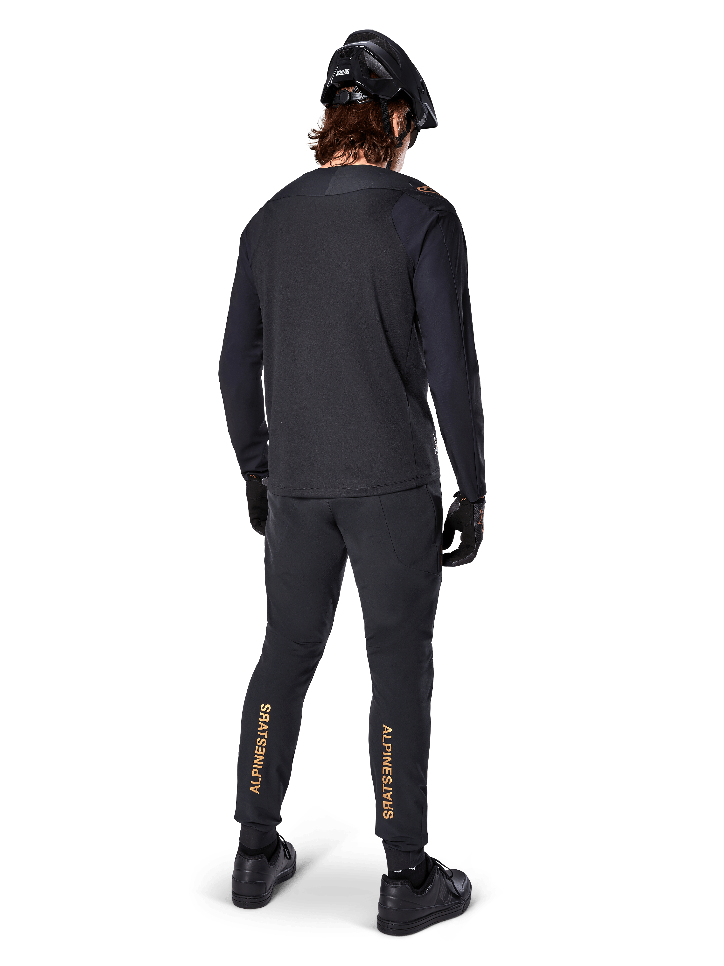 Set bagfra af en mountainbike-rytter iført Alpinestars A-Aria Polartec Switch langærmet jersey i sort, kombineret med sorte MTB-bukser med tan-farvet Alpinestars-branding på læggene, sort hjelm og handsker, professionelt cykeludstyr til off-road stier