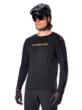 Alpinestars A-Aria Polartec Switch langærmet trøje, sort med guldlogo, MTB mountainbike-beklædning, iført sort hjelm og solbriller, med laserskåret ventilation på ærmerne og svedtransporterende Polartec-stof til terrænkørsel