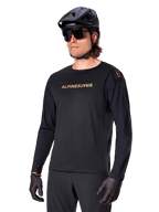 Alpinestars A-Aria Polartec Switch langærmet trøje, sort med guldlogo, MTB mountainbike-beklædning, iført sort hjelm og solbriller, med laserskåret ventilation på ærmerne og svedtransporterende Polartec-stof til terrænkørsel