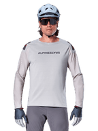 Alpinestars A-Aria Polartec Switch langærmet jersey, lysegrå mountainbike-jersey med laserperforeringer på ærmerne og brandnavn på brystet, båret med lyseblå hjelm, solbriller og grå handsker