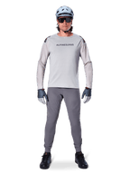 Alpinestars A-Aria Polartec Switch Jersey, langærmet, lysegrå, båret af en mandlig mountainbikerytter med mørkegrå MTB-bukser, lyseblå hjelm og solbriller, med branding på brystet og laserperforeret ventilation på ærmerne, komplet off-road køreudstyr.