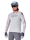 Alpinestars A-Aria Polartec Switch langærmet trøje, lysegrå cykeltrøje med sort brystlogo og røde skulderdetaljer, laserperforerede ærmer for ventilation, vist med lyseblå hjelm, solbriller og grå bukser, performance mountainbike-tøj til trail-kørsel