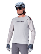 Alpinestars A-Aria Polartec Switch langærmet trøje, lysegrå cykeltrøje med sort brystlogo og røde skulderdetaljer, laserperforerede ærmer for ventilation, vist med lyseblå hjelm, solbriller og grå bukser, performance mountainbike-tøj til trail-kørsel