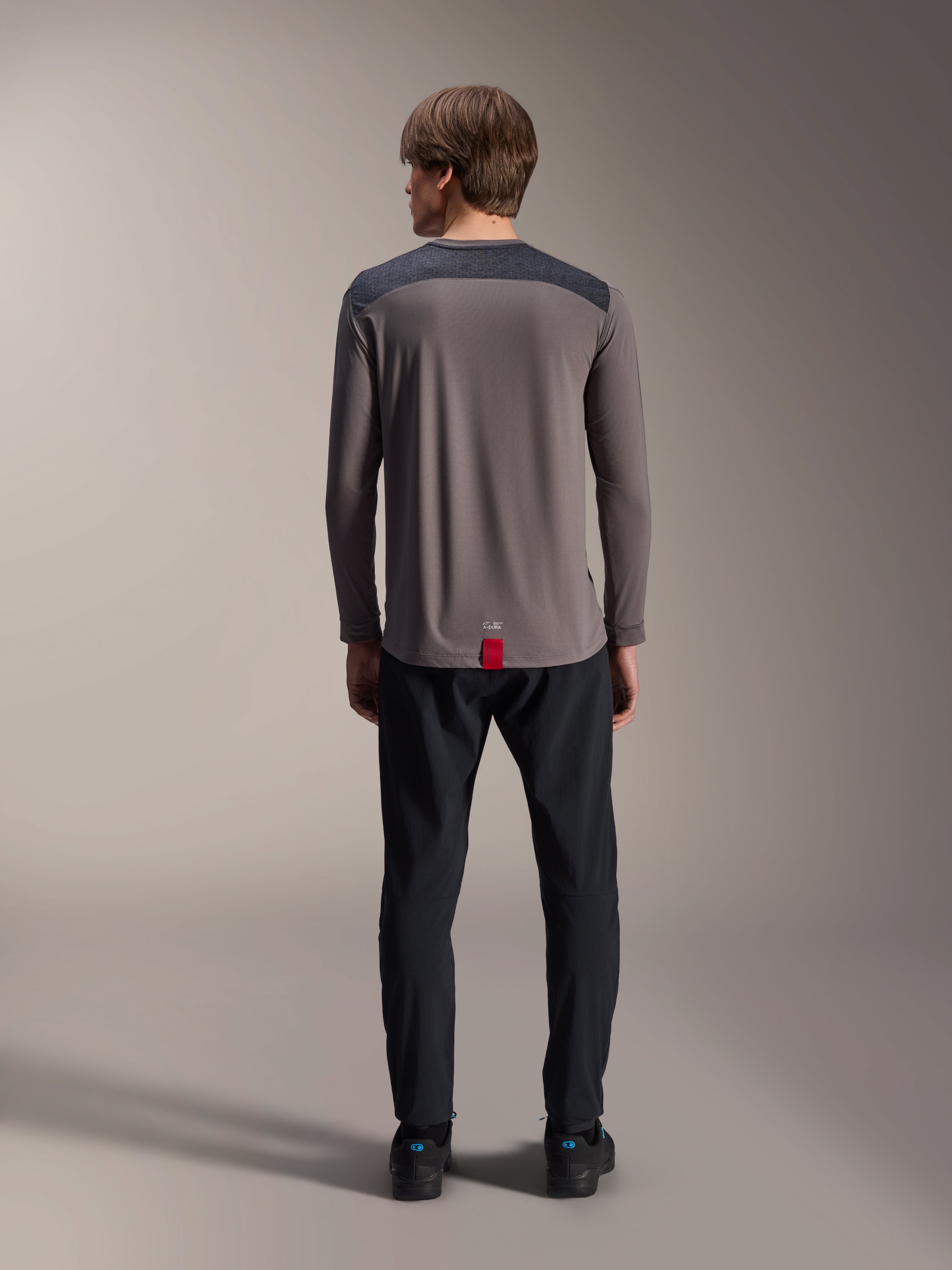 Alpinestars A-Dura Insert Astar langærmet jersey i mørkegrå, set bagfra, med koksgrå skulderpaneler og et rødt mærke ved sømmen, kombineret med sorte bukser og sko, mountainbike performance-beklædning