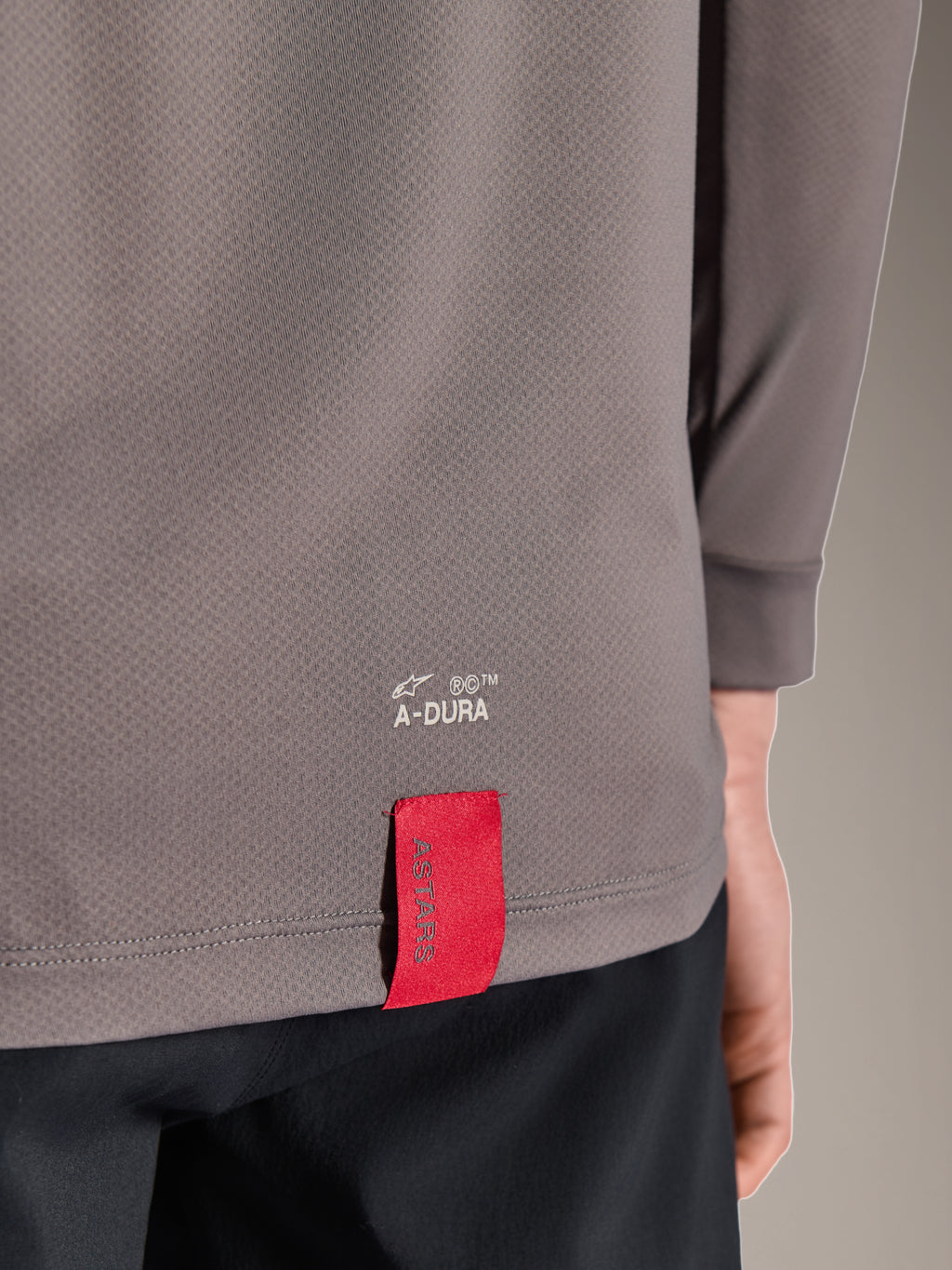 Alpinestars A-Dura Insert Astar Jersey, langærmet MTB-trøje, mørkegrå, nærbillede af detalje på den nederste kant på ryggen med hvid A-DURA-branding og et karakteristisk rødt ASTARS-stofmærke, tekstureret performance-materiale