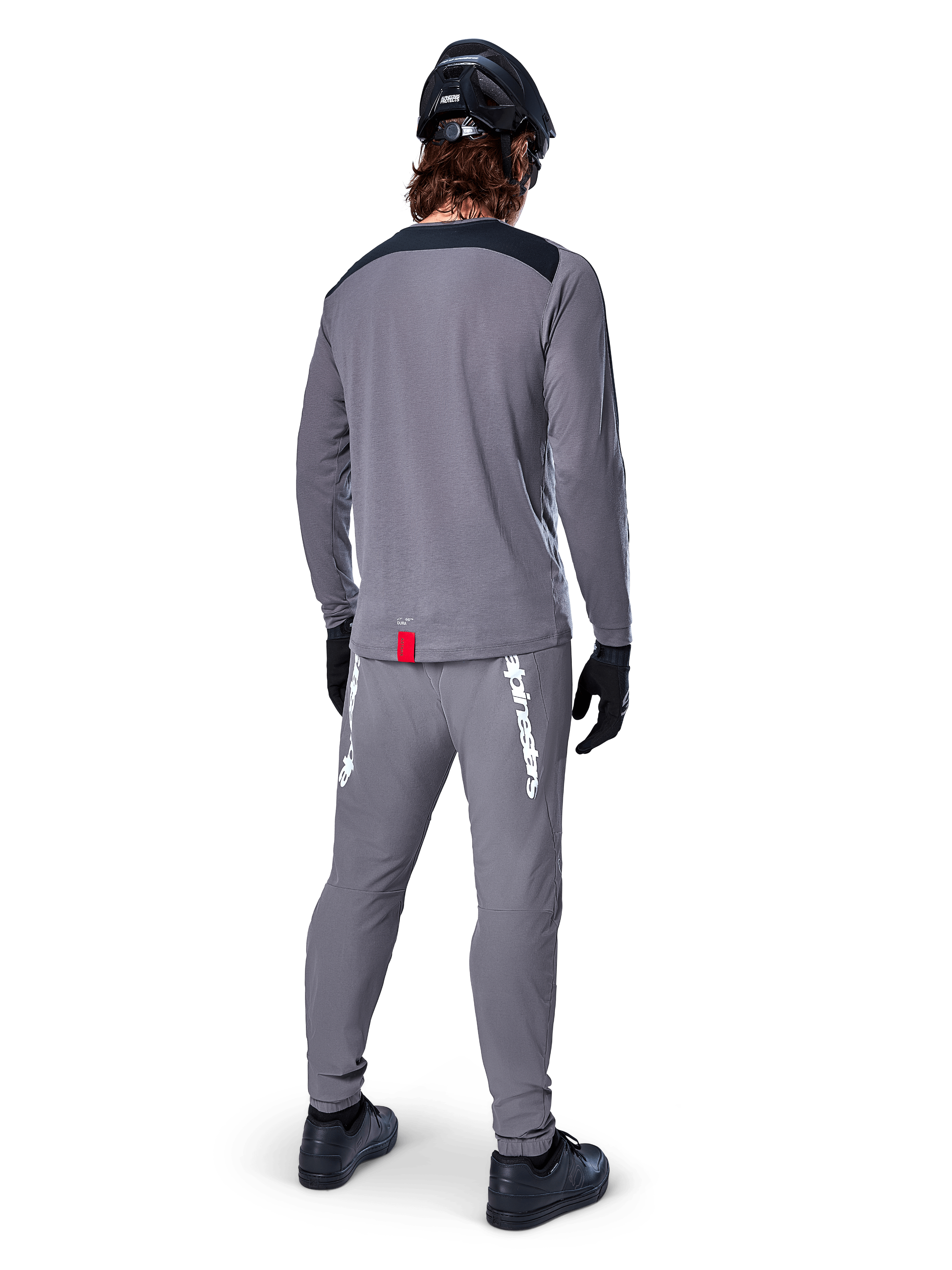 Alpinestars A-Dura Dri Astar langærmet jersey, mørkegrå, set bagfra af rytter iført matchende bukser og sort hjelm, har sorte skulderpaneler og et lille rødt mærke ved kanten, teknisk off-road cykeludstyr