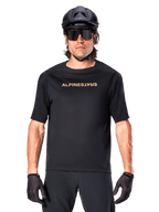 Mountainbiker iført Alpinestars A-Aria Polartec Switch kortærmet trøje i sort med guldtonet brystlogo, sort MTB-hjelm, mørke performance-solbriller og matchende handsker, off-road cykeludstyr.