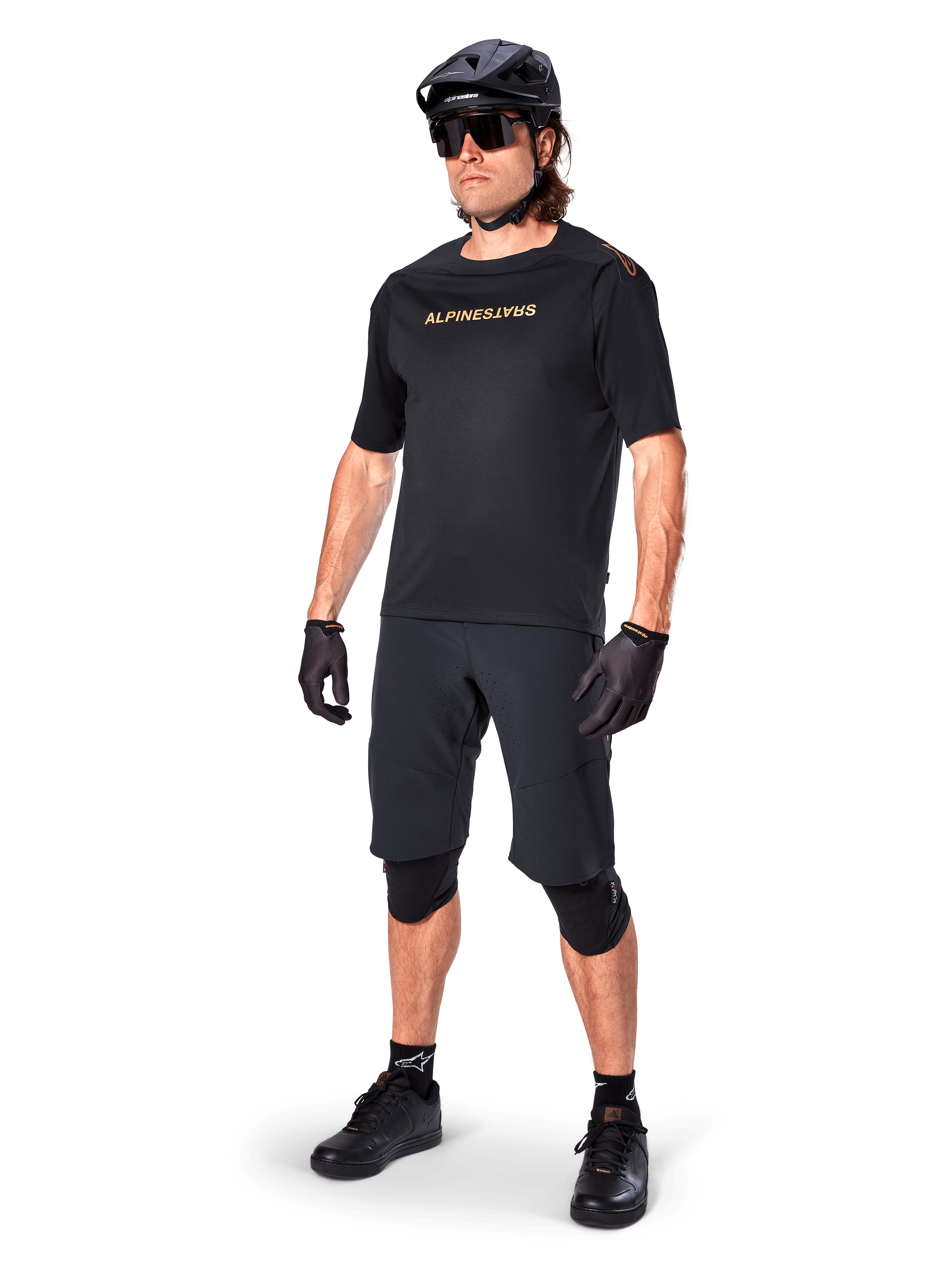Alpinestars A-Aria Polartec Switch kortærmet trøje, sort med spejlvendt guld-branding på brystet, båret af en rytter med sorte cykelshorts, hjelm og knæbeskyttere, teknisk MTB-udstyr til terrænkørsel