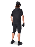 Mandlig mountainbikerytter set bagfra iført Alpinestars A-Aria Polartec Switch Jersey i sort, kortærmet MTB-trøje, kombineret med sorte mountainbike-shorts, sort hjelm og handsker, koordineret performance-outfit til terrænkørsel på stier.