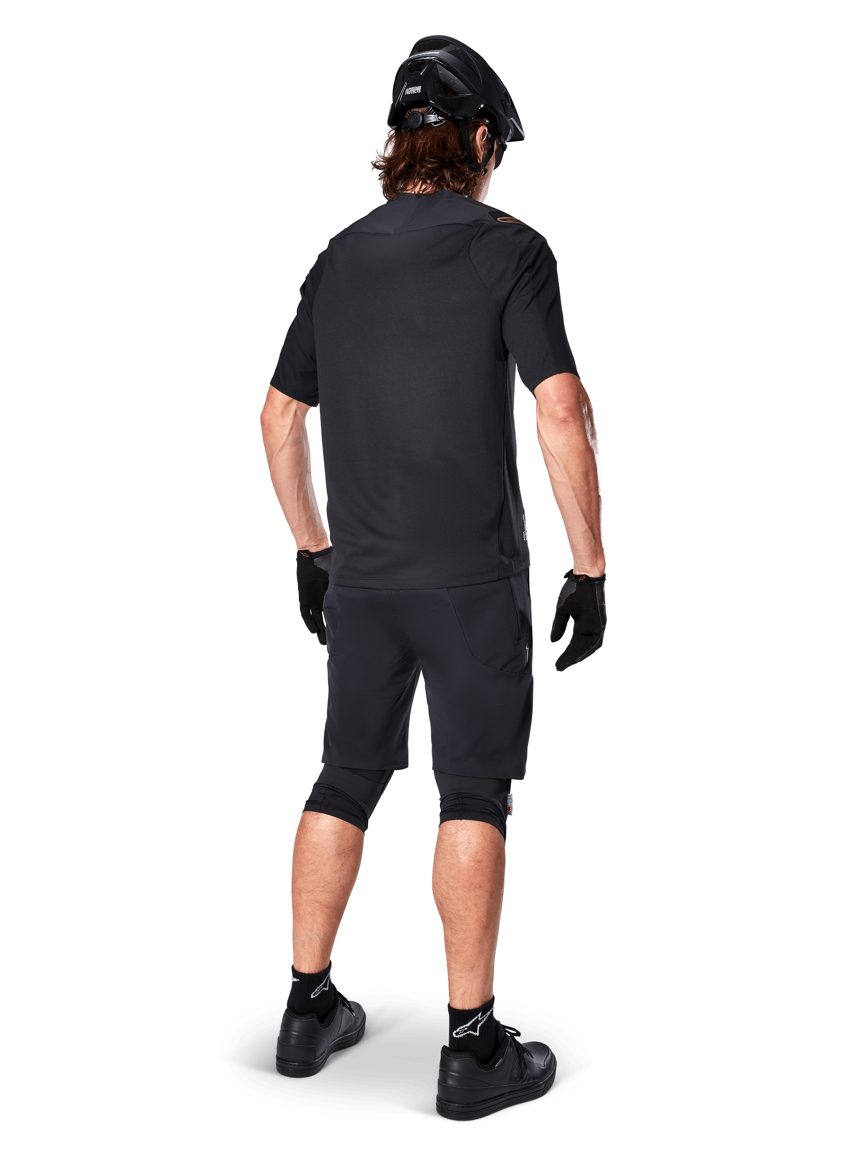 Mandlig mountainbikerytter set bagfra iført Alpinestars A-Aria Polartec Switch Jersey i sort, kortærmet MTB-trøje, kombineret med sorte mountainbike-shorts, sort hjelm og handsker, koordineret performance-outfit til terrænkørsel på stier.