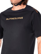 Alpinestars A-Aria Polartec Switch trøje, kortærmet MTB-trøje, sort, guld Alpinestars-skrift på brystet og logo på skulderen, nærbillede af overkroppen, der fremhæver teknisk stof og performance-pasform