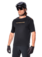Alpinestars A-Aria Polartec Switch kortærmet cykeltrøje, sort mountainbike-trøje med guld Alpinestars-branding, sort hjelm, solbriller og handsker, teknisk MTB-beklædning