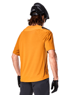 Alpinestars A-Aria Polartec Switch trøje, kortærmet MTB-trøje, mørkeguld, set bagfra med performance fit, båret med sort hjelm og grå shorts, slidstærkt og åndbart udstyr til trail-kørsel