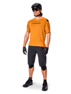 Mandlig rytter iført Alpinestars A-Aria Polartec Switch trøje i Dark Gold, kortærmet MTB-trøje, kombineret med sorte cykelshorts, knæbeskyttere, sort hjelm og handsker, professionelt mountainbike-outfit til trail-kørsel