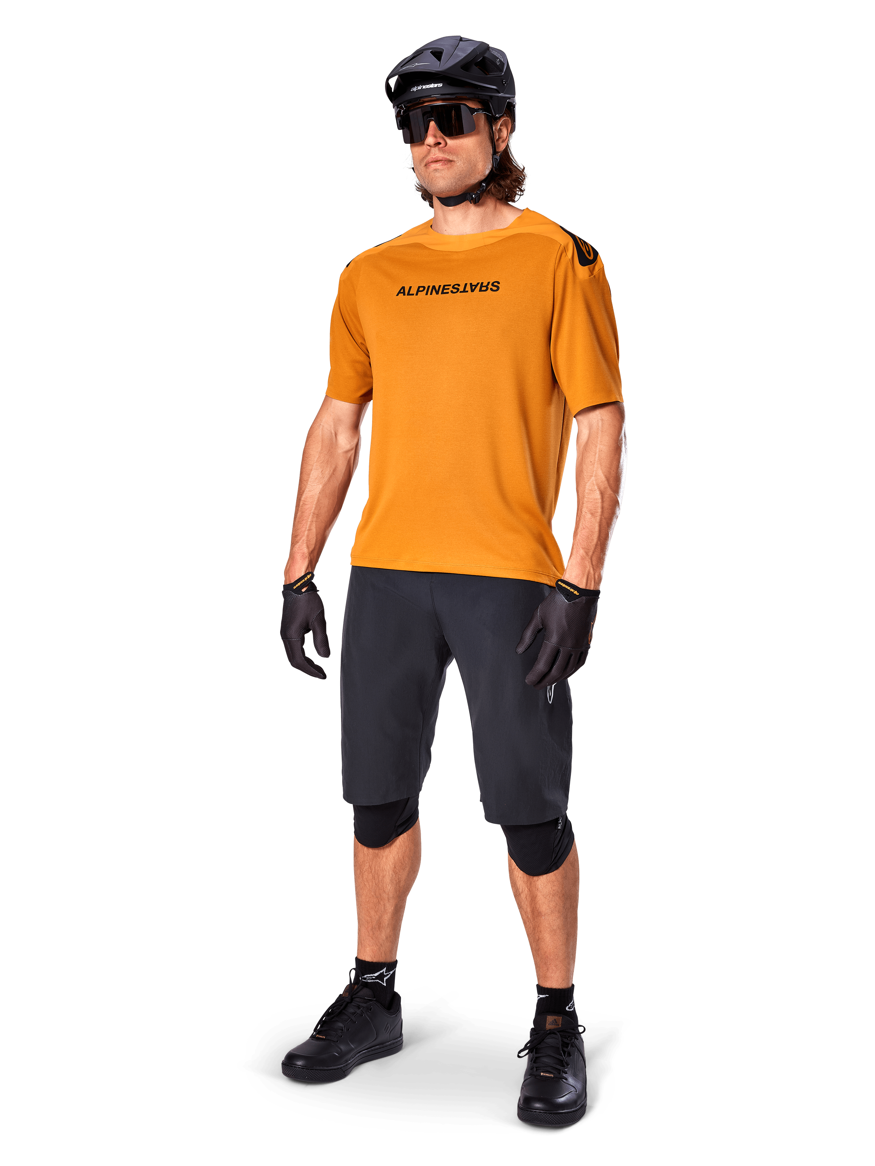 Mandlig rytter iført Alpinestars A-Aria Polartec Switch trøje i Dark Gold, kortærmet MTB-trøje, kombineret med sorte cykelshorts, knæbeskyttere, sort hjelm og handsker, professionelt mountainbike-outfit til trail-kørsel