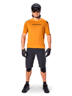 Mandlig cyklist iført Alpinestars A-Aria Polartec Switch kortærmet trøje i Dark Gold, kombineret med sorte MTB-shorts, hjelm, solbriller og handsker, professionelt mountainbike-udstyr til trailkørsel