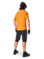 Alpinestars A-Aria Polartec Switch-trøje, kortærmet MTB-trøje, Dark Gold, set bagfra af model iført mørkegrå mountainbike-shorts, sort hjelm og handsker, teknisk cykeltøj til off-road stier