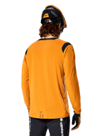 Alpinestars A-Dura Dri Wool langærmet jersey i Dark Gold, set bagfra af en rytter iført mountainbikehjelm og handsker, med sorte mesh-paneler på skuldrene og et længere rygstykke, teknisk cykeltøj til trailkørsel