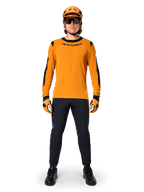 Mountainbiker iført Alpinestars A-Dura Dri Wool langærmet trøje, mørk guld med sorte detaljer på hals og ærmer, kombineret med sorte bukser, matchende guldhjelm, handsker og sorte solbriller, professionelt mountainbike-udstyr