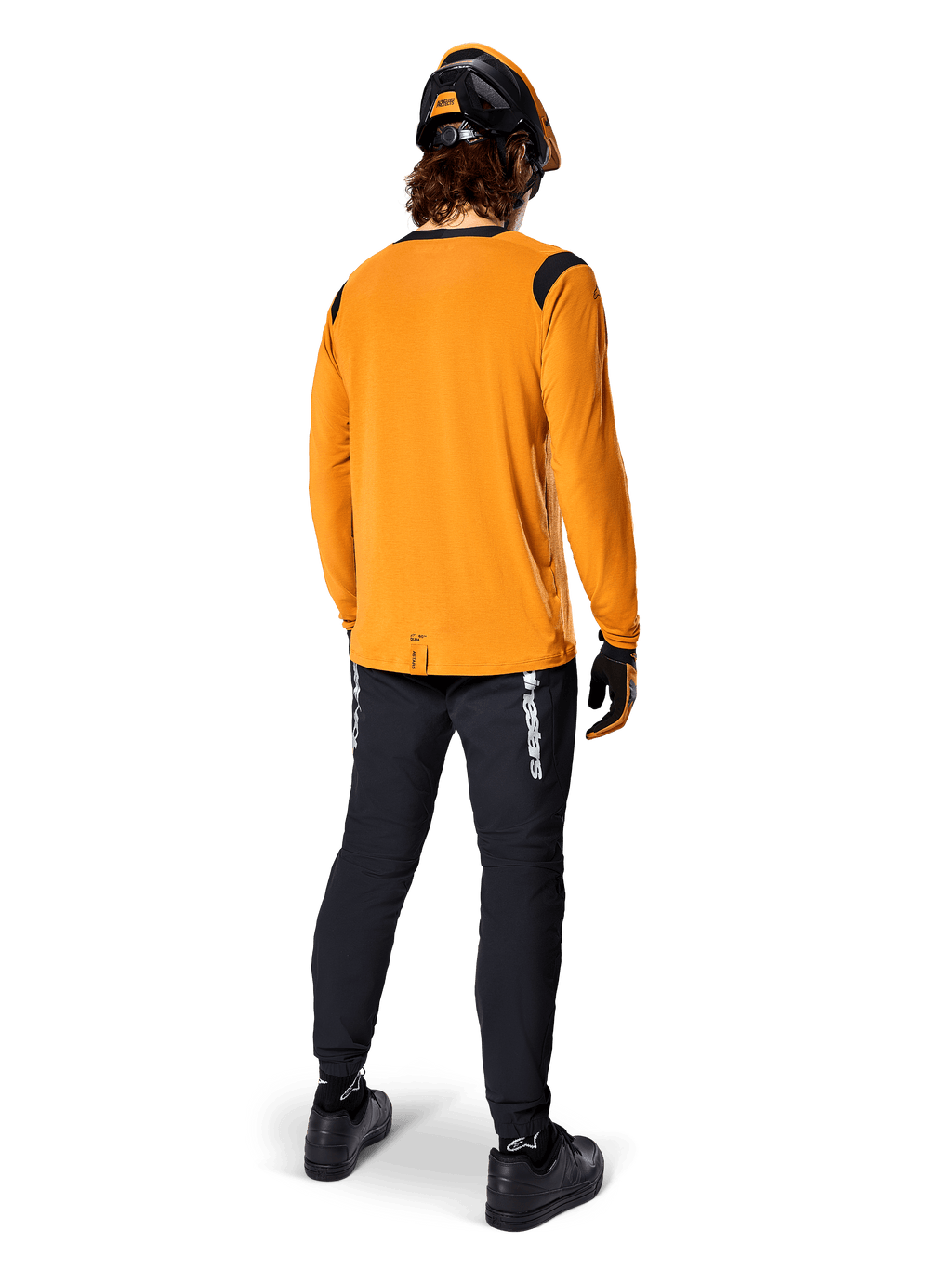 Mountainbiker set bagfra iført Alpinestars A-Dura Dri Wool langærmet trøje i Dark Gold, sorte tekniske bukser med hvidt branding og en matchende sort og orange hjelm, præstationsorienteret mountainbike-beklædning.