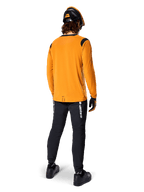 Mountainbiker set bagfra iført Alpinestars A-Dura Dri Wool langærmet trøje i Dark Gold, sorte tekniske bukser med hvidt branding og en matchende sort og orange hjelm, præstationsorienteret mountainbike-beklædning.