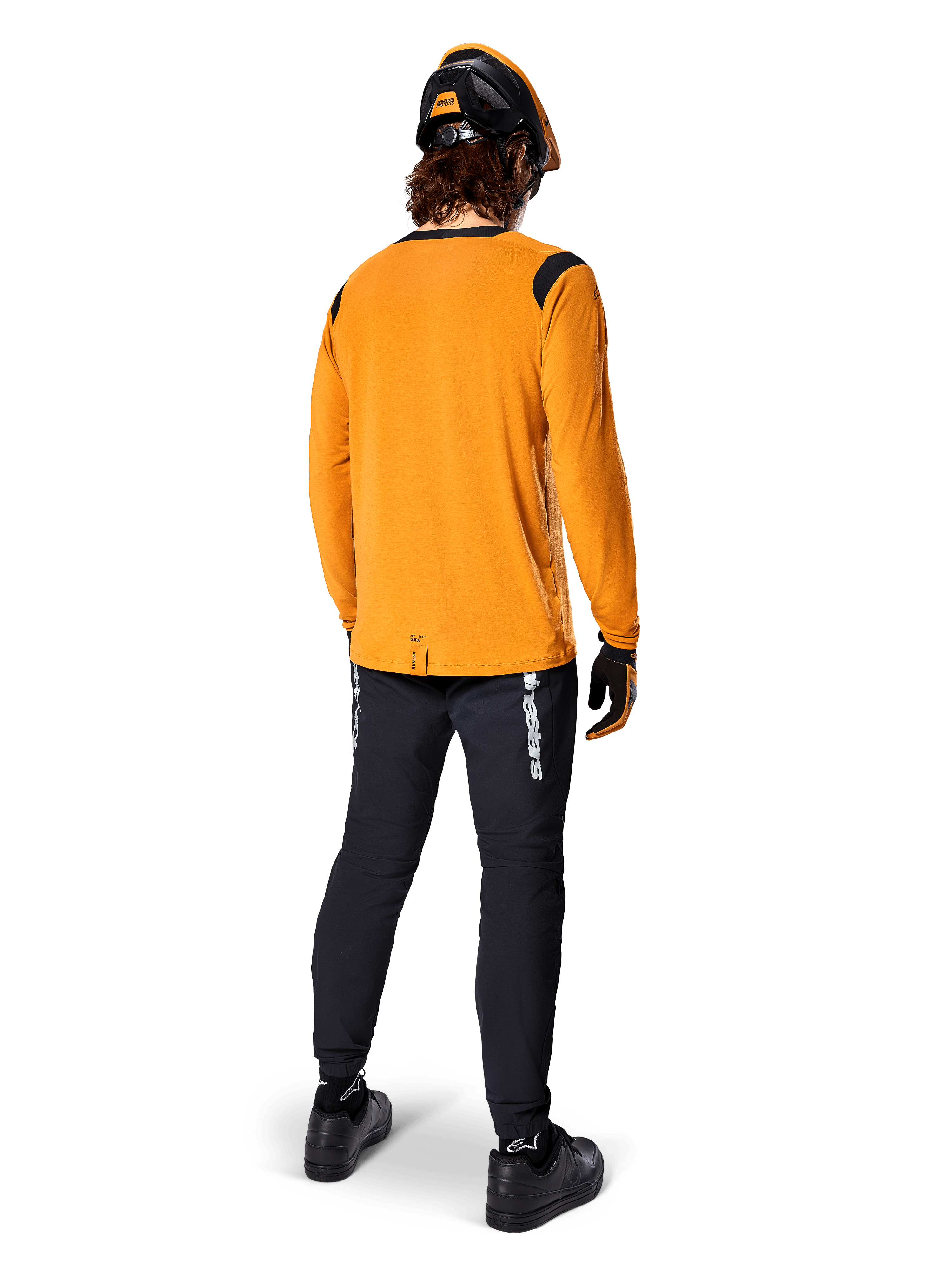Mountainbiker set bagfra iført Alpinestars A-Dura Dri Wool langærmet trøje i Dark Gold, sorte tekniske bukser med hvidt branding og en matchende sort og orange hjelm, præstationsorienteret mountainbike-beklædning.