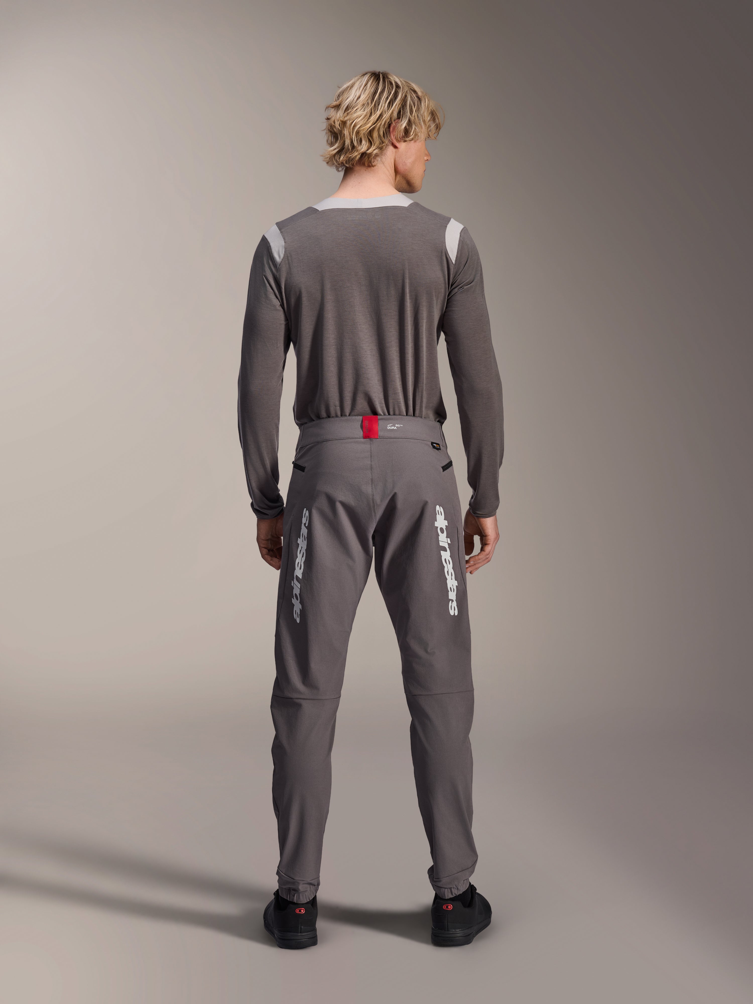 Model set bagfra iført Alpinestars A-Dura Elite bukser i mørkegrå og en matchende grå langærmet trøje, med hvide Alpinestars-logoer på benene, en rød detalje på linningen og sorte sko, professionelt MTB-udstyr.
