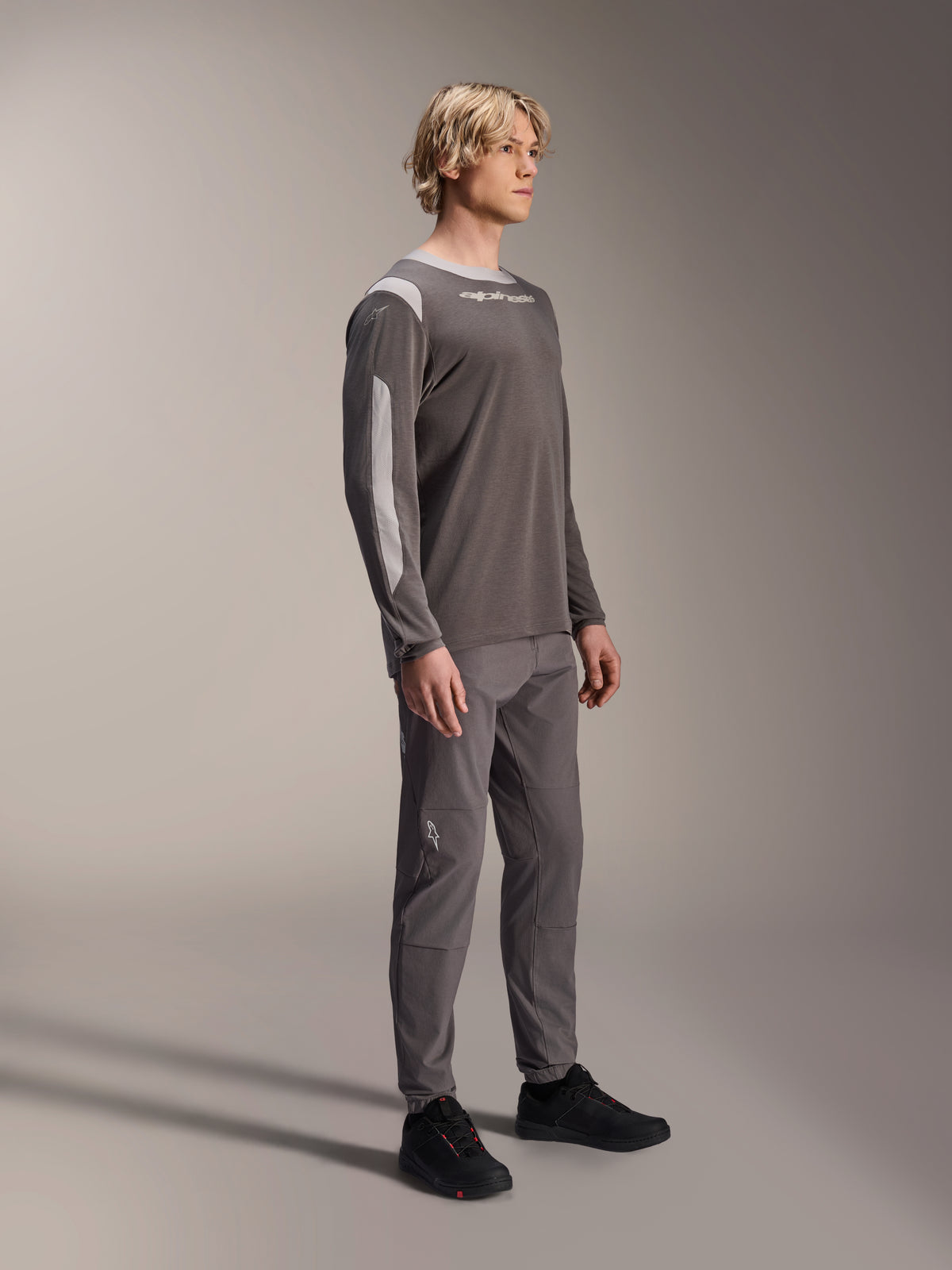 Mandlig model iført Alpinestars A-Dura Dri Wool langærmet trøje i mørkegrå, kombineret med matchende grå bukser og sorte sko, med lysegrå mesh-paneler på skuldre og ærmer, teknisk MTB-udstyr til terrænkørsel