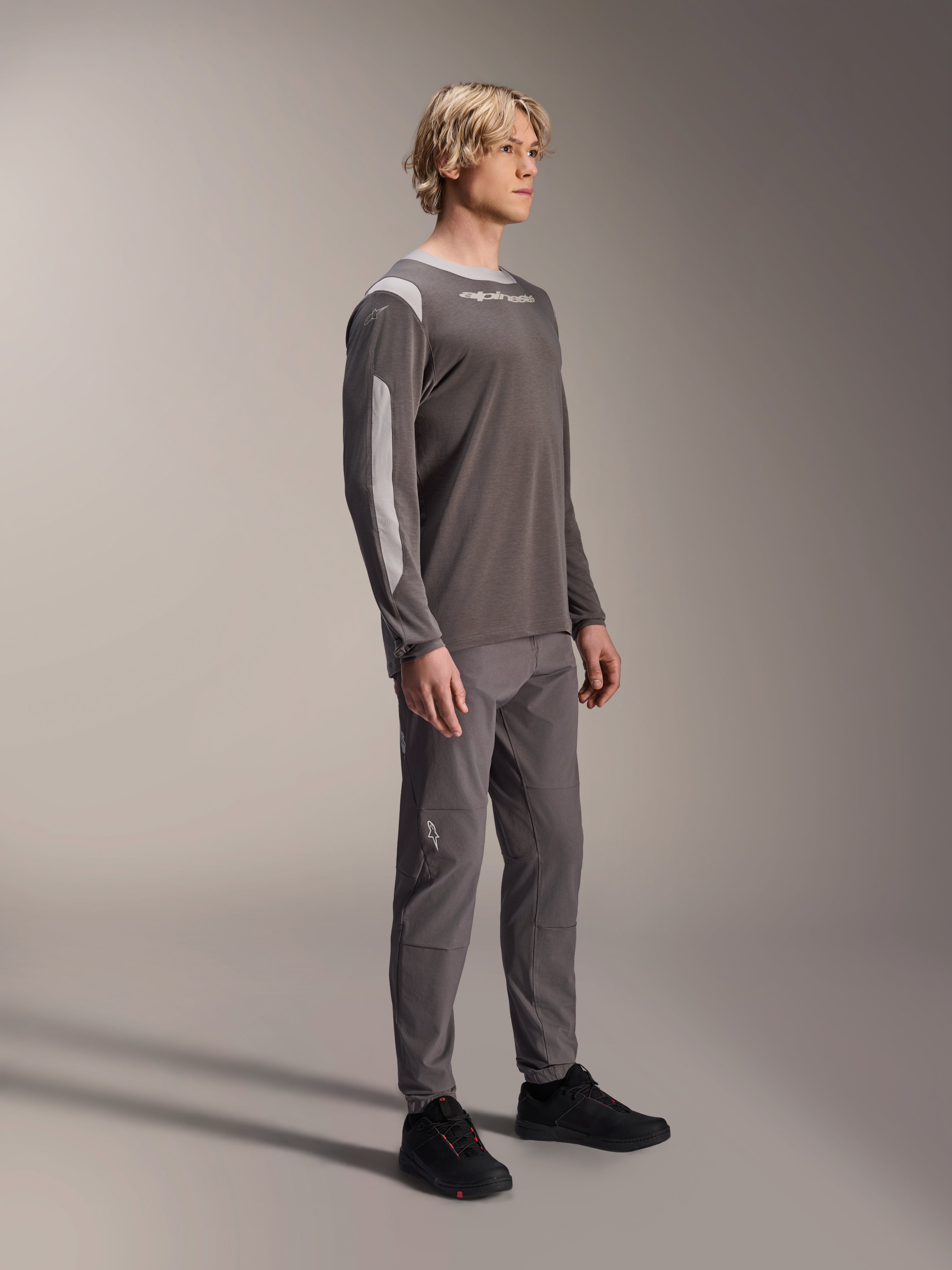 Mandlig model iført Alpinestars A-Dura Dri Wool langærmet trøje i mørkegrå, kombineret med matchende grå bukser og sorte sko, med lysegrå mesh-paneler på skuldre og ærmer, teknisk MTB-udstyr til terrænkørsel